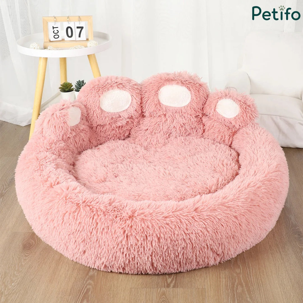An empty pink fluffy paw-shaped dog bed displayed in a bright room with a small side table.