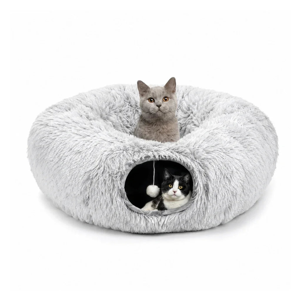 A gray cat sitting inside a plush donut bed with a black and white cat peeking out of the tunnel entrance